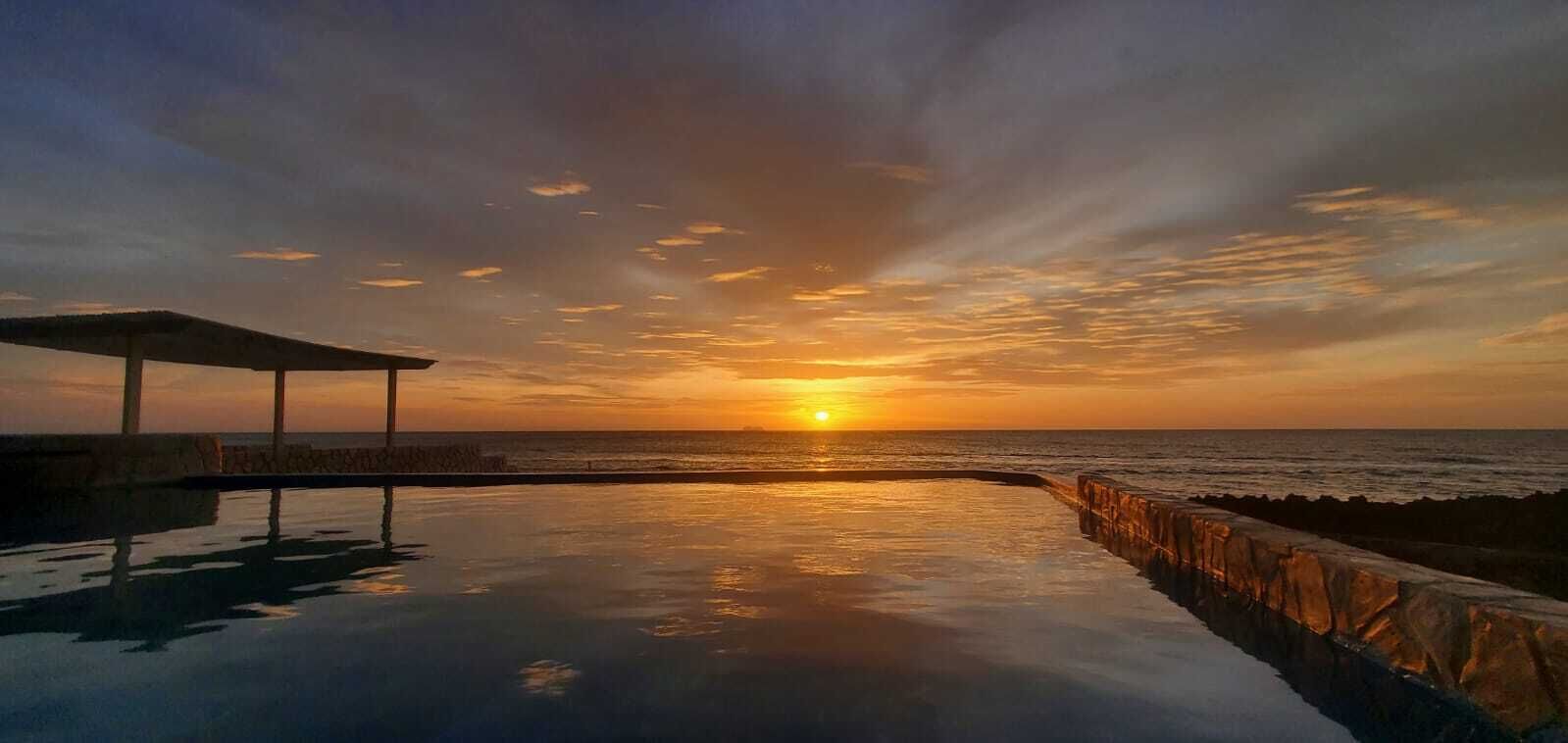 Vista de un atardecer desde una piscina infinita con barandilla de piedra y zona de estar cubierta.