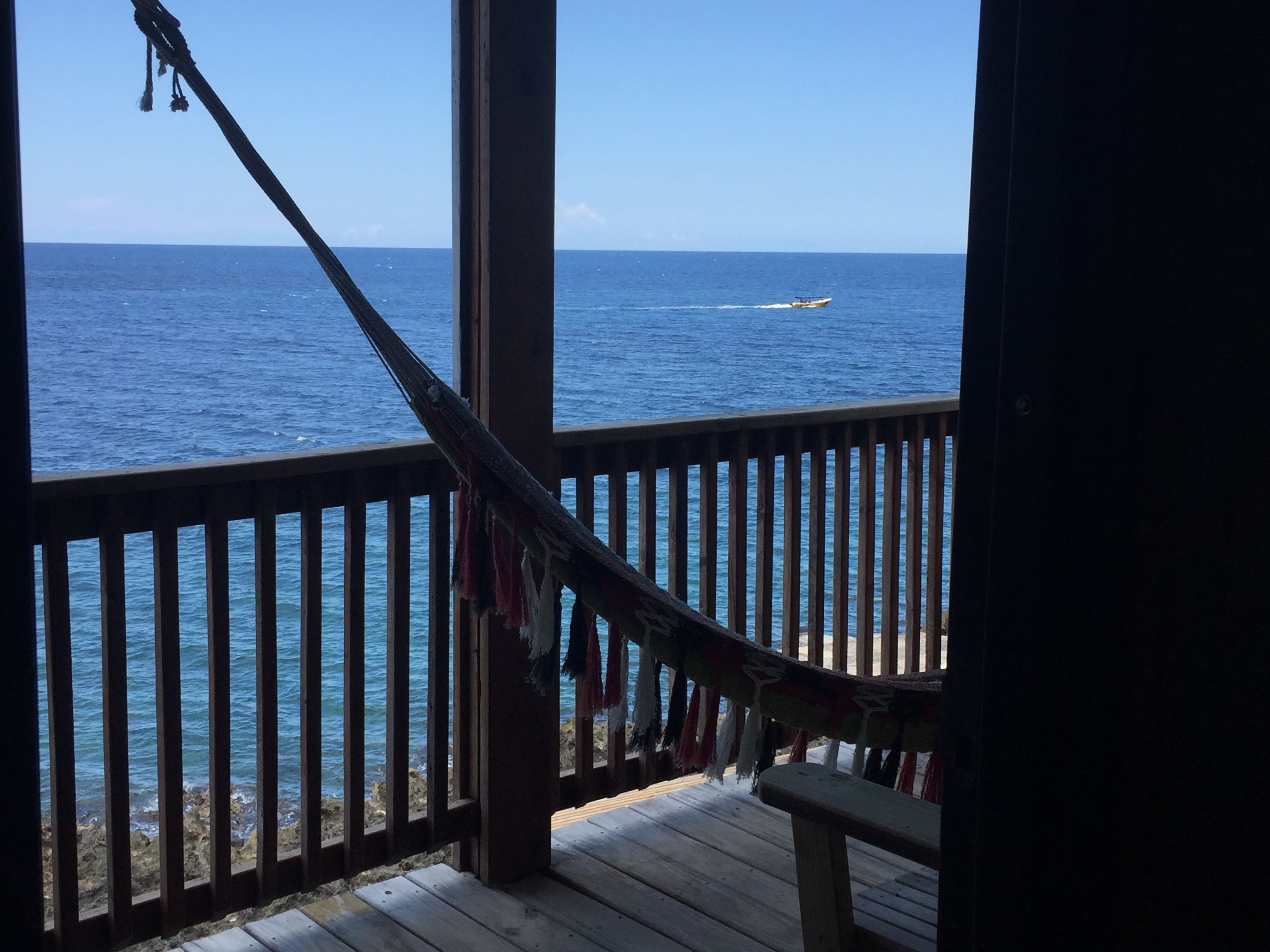 A wooden deck with a hammock and clothes hanging, overlooking a calm sea with a boat in the distance.