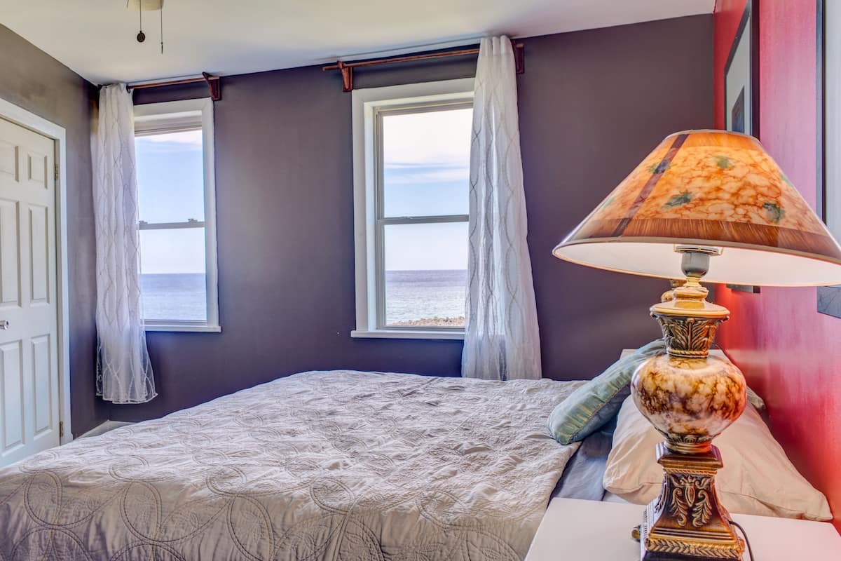 A bedroom with a bed, two windows with curtains, a lamp, and a view of the sea.
