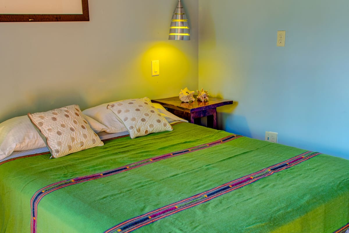 A neatly made bed with a green bedspread and white pillows, a wooden nightstand with a lamp, and a framed picture on the wall.