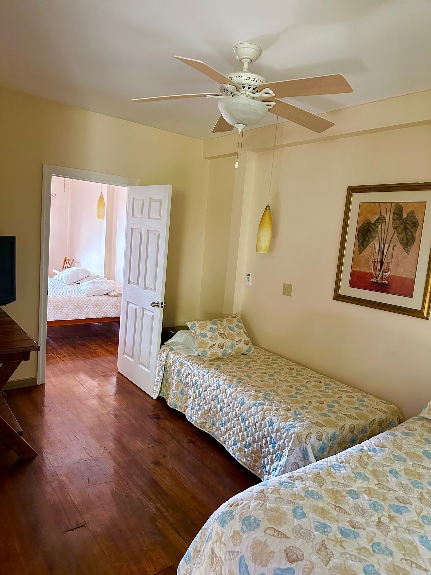A room with two beds, a ceiling fan, a door leading to another room, and a framed picture on the wall.