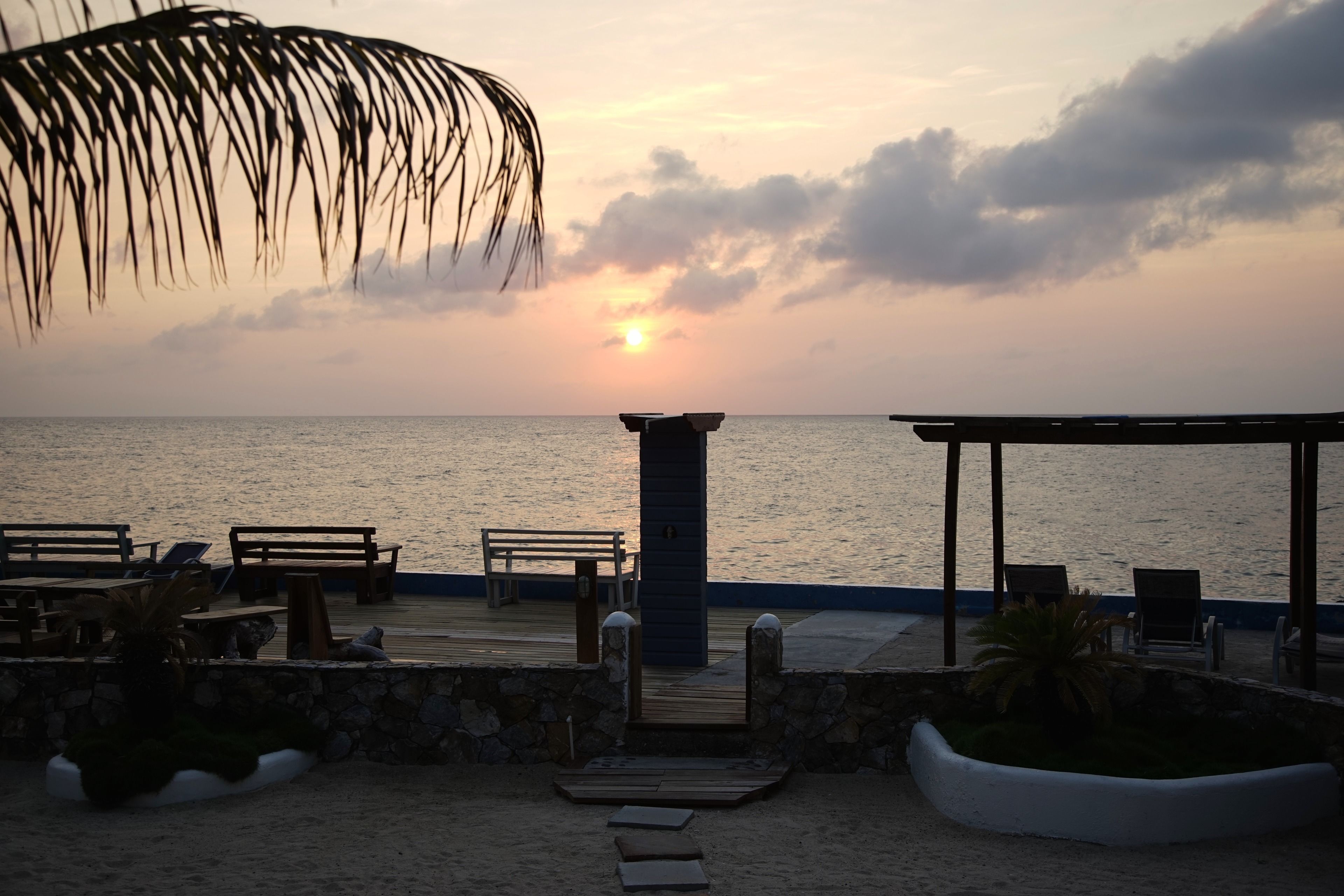 Un patio frente a la playa con bancos y mesas de madera, un muro de piedra y vistas al océano al atardecer.