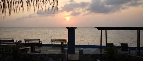 Un patio frente a la playa con bancos y mesas de madera, un muro de piedra y vistas al océano al atardecer.
