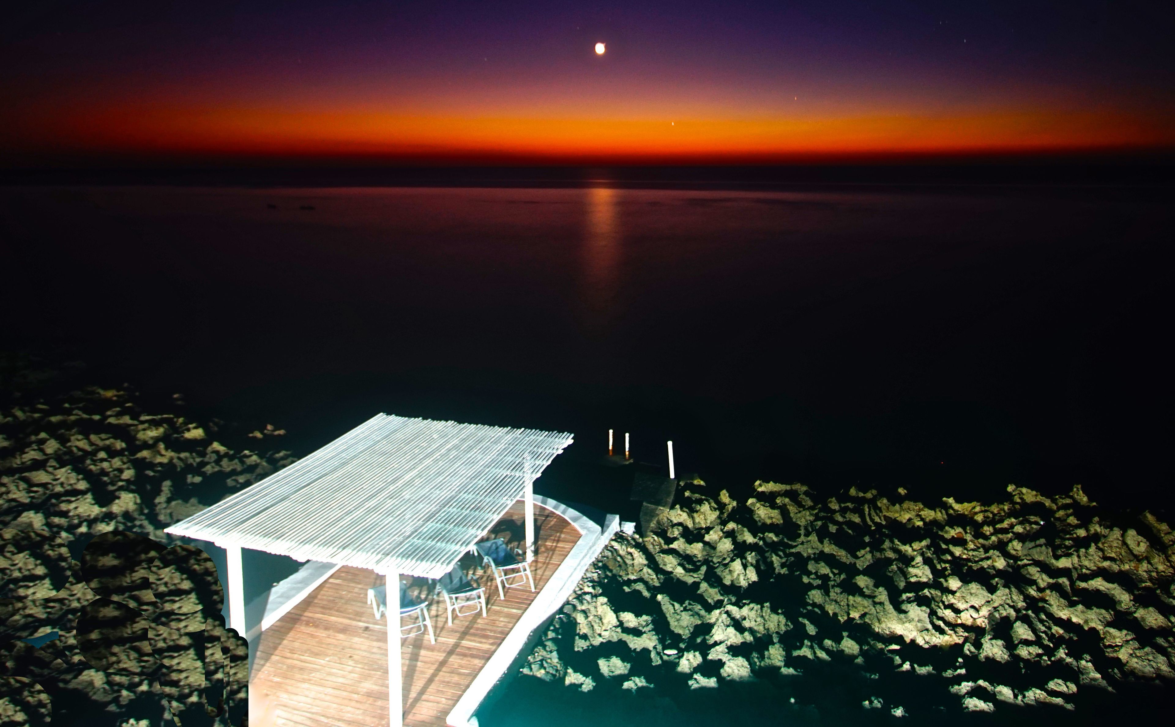 Un muelle de madera se extiende sobre aguas tranquilas de noche, bajo un cielo despejado con una luna visible.
