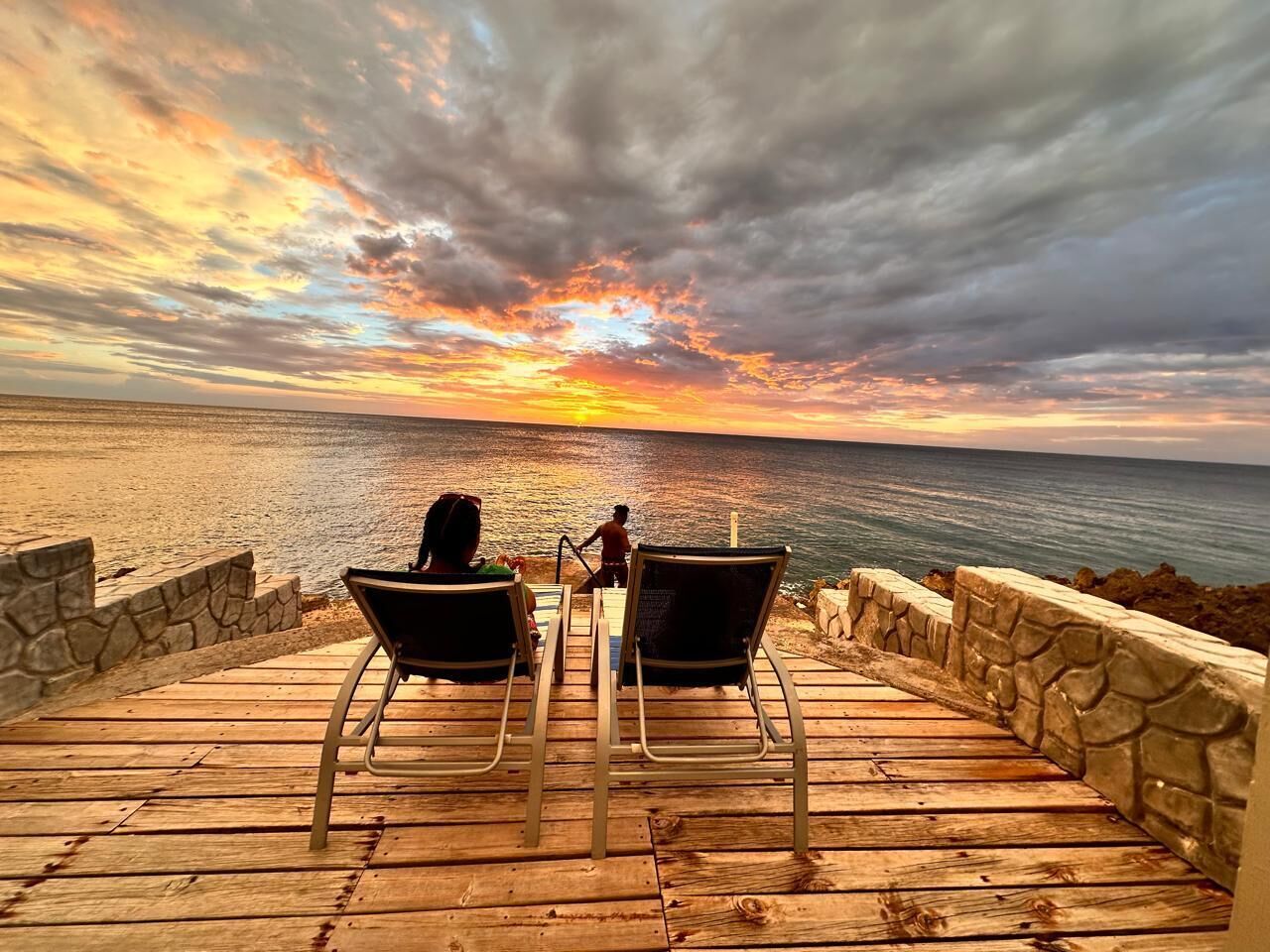 A wooden deck with two lounge chairs facing a beach, overlooking the ocean.