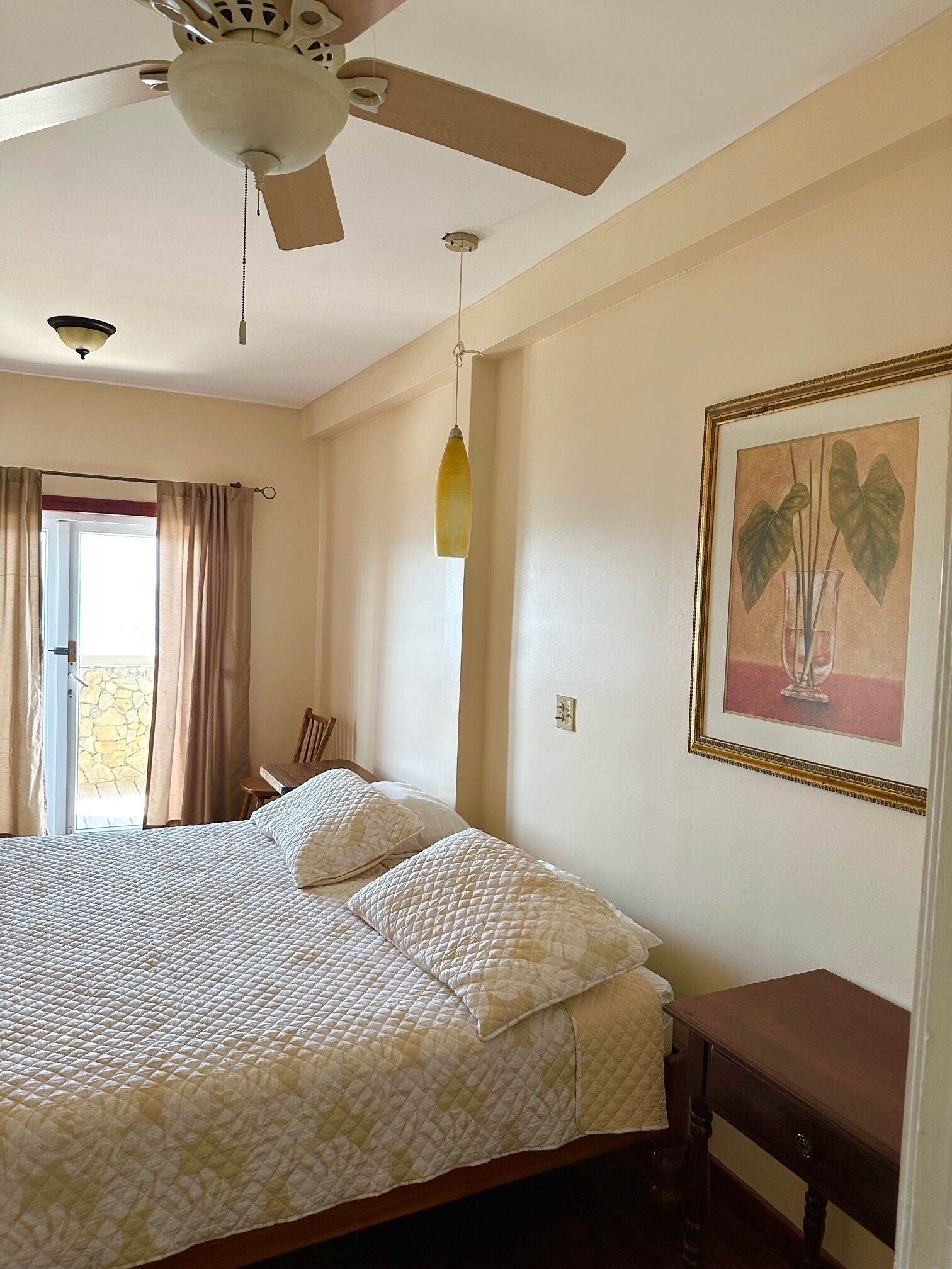 Un dormitorio con cama, ventilador de techo, mesita de noche, una silla, ventana con cortinas e imagen enmarcada en la pared.
