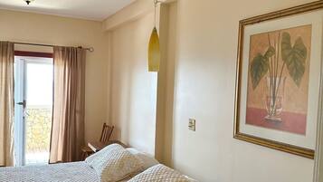 Un dormitorio con cama, ventilador de techo, mesita de noche, una silla, ventana con cortinas e imagen enmarcada en la pared.