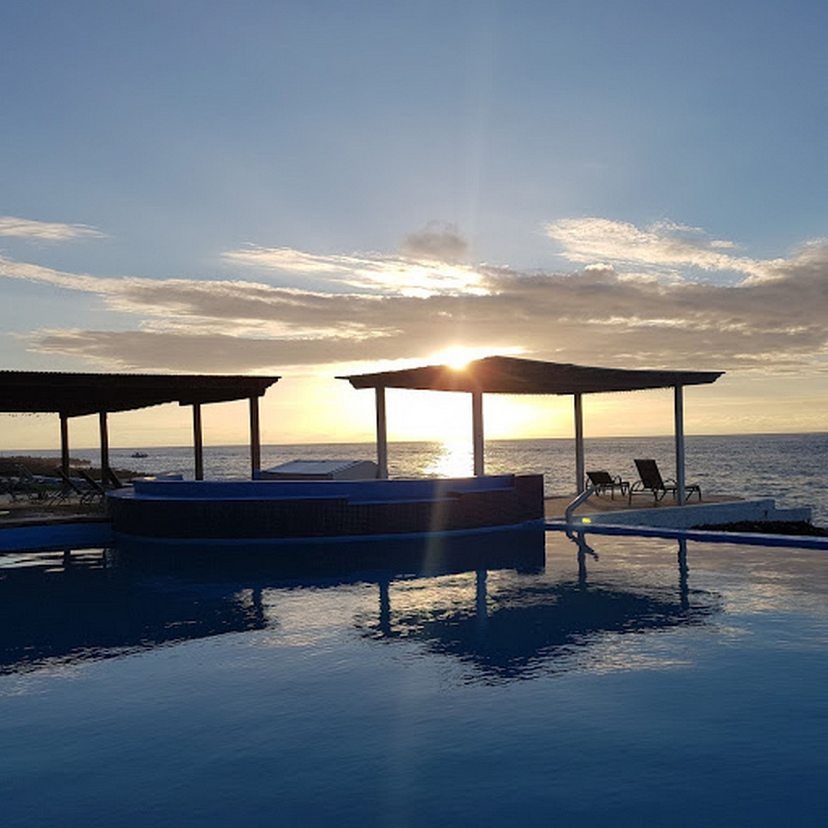 Piscina con vista al atardecer, sillas de descanso y un área de estar cubierta.