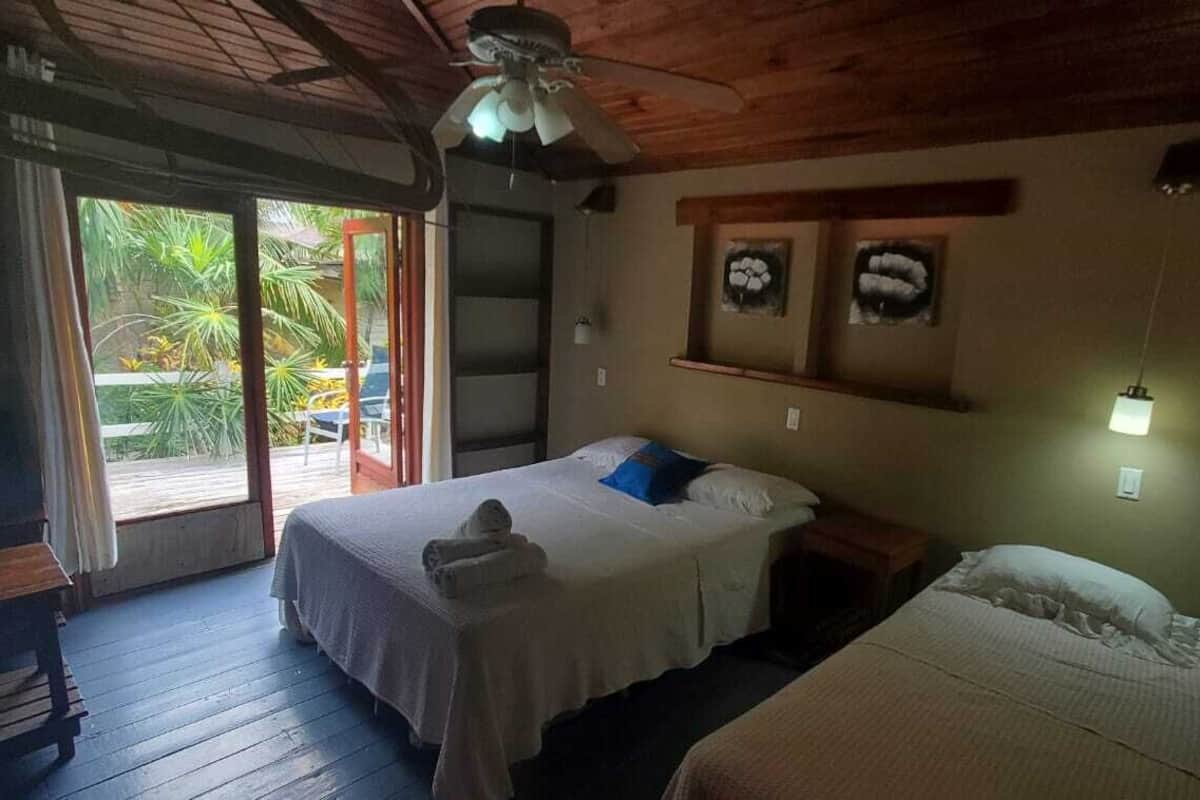 A room with two beds, a ceiling fan, a small table, and a window with a view of greenery outside.
