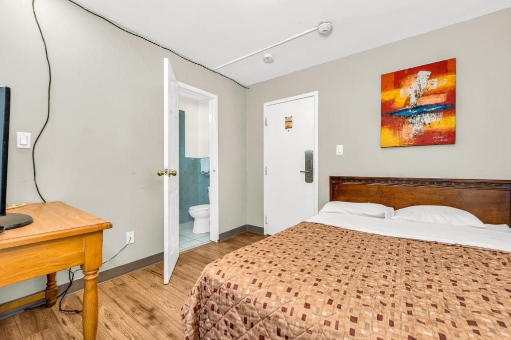 A hotel room with a bed, a desk, a painting on the wall, and a bathroom visible through an open door.
