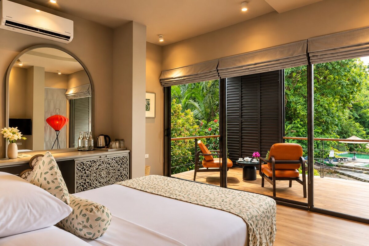 Habitación de hotel con cama, escritorio con espejo, vista a un balcón con sillas y un ventanal grande.
