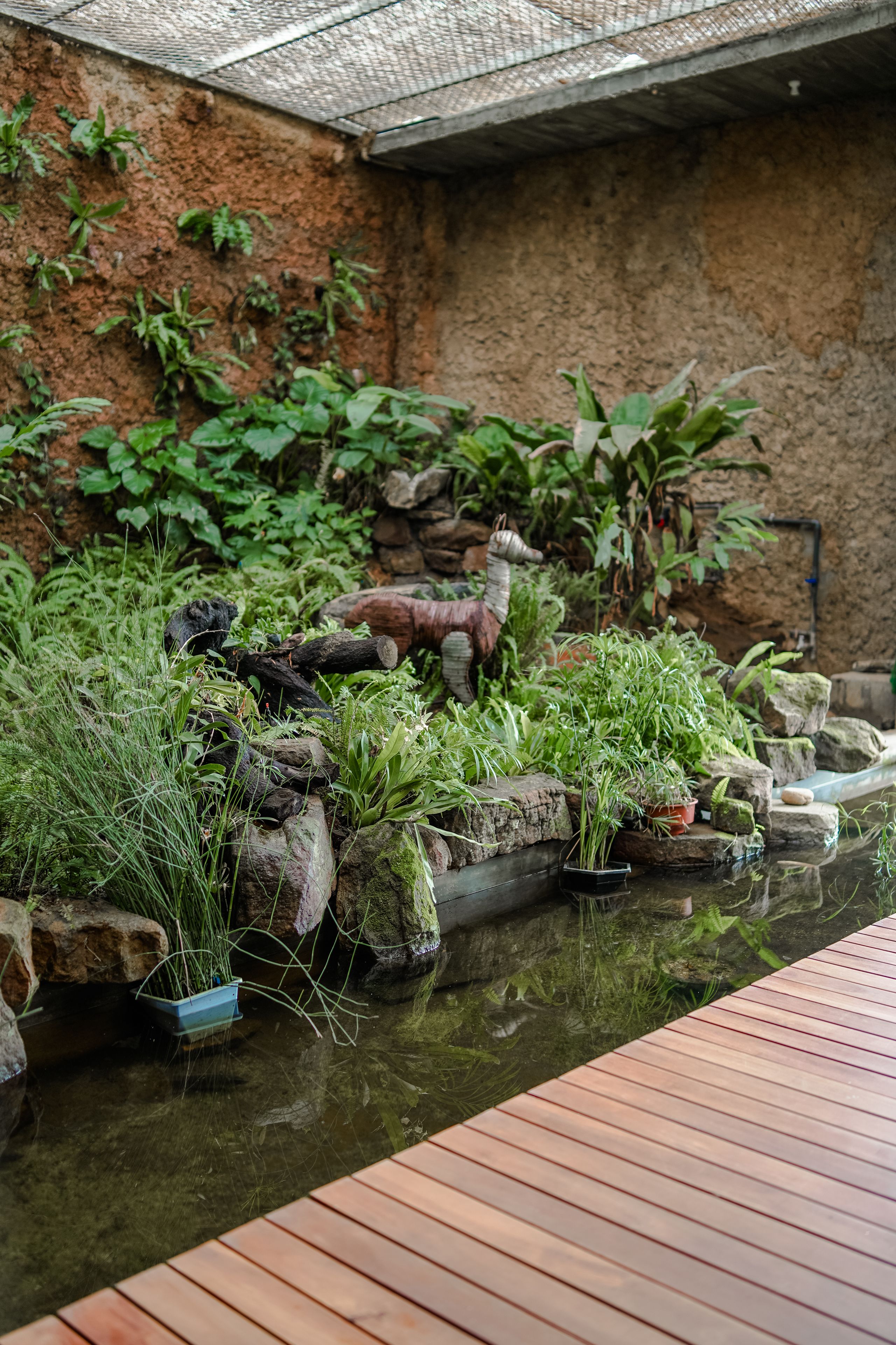 A serene indoor garden with a wooden deck, various plants, and a small water feature.