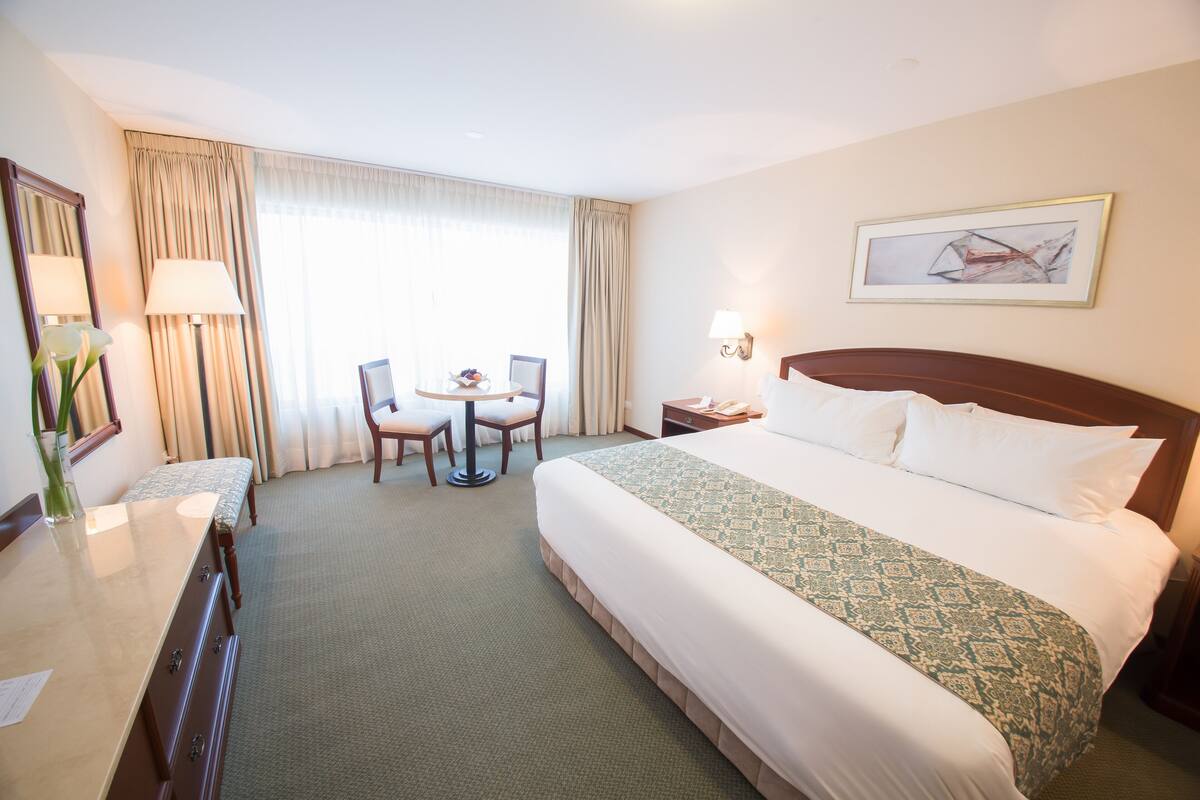 A hotel room with a large bed, a desk with a lamp, a chair, a small table, and a window with curtains.