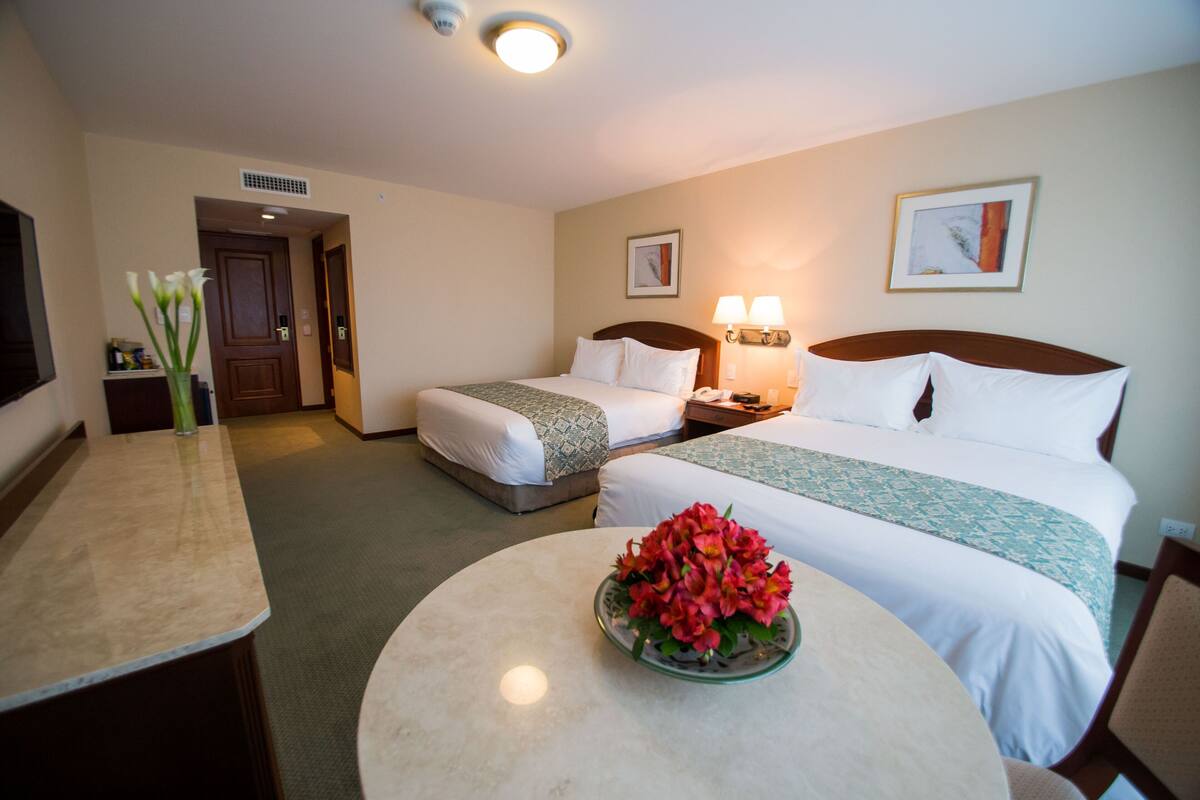 A hotel room with two beds, a round table with a vase of flowers, a desk, and a television.