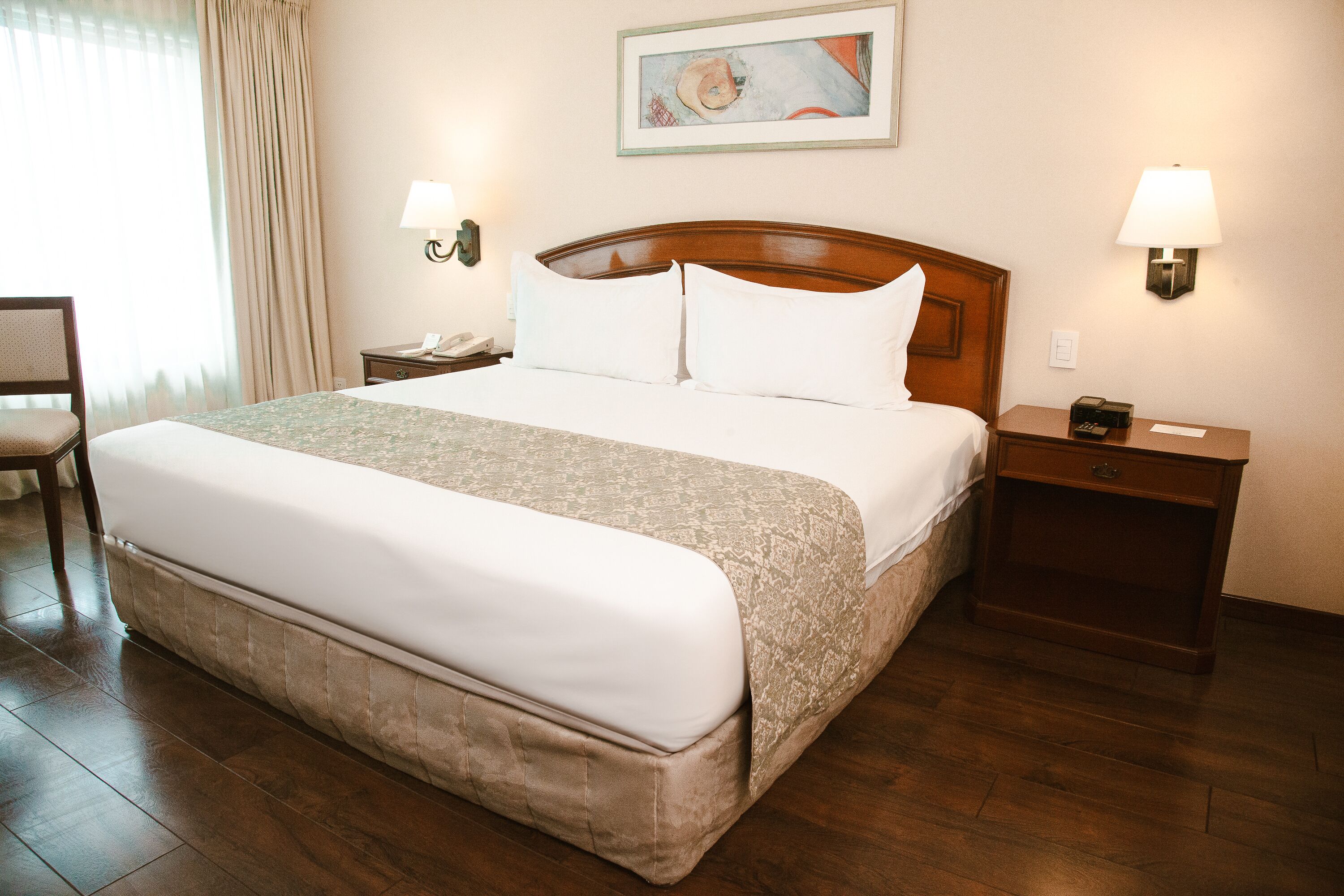 Una habitación de hotel con una cama grande, dos mesitas de noche, una silla y un cuadro en la pared.