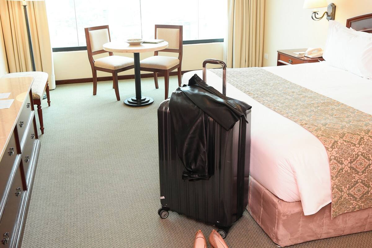 A hotel room with a bed, a desk, a chair, a table, a lamp, a suitcase, and a pair of shoes.