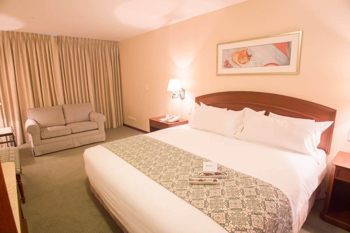 A hotel room with a large bed, a sofa, a nightstand with a lamp, and a painting on the wall.