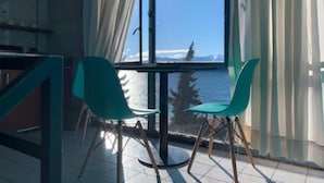 Una habitación con un ventanal amplio que ofrece vistas al mar y a las montañas. Hay dos sillones verdes y una mesa en la habitación.