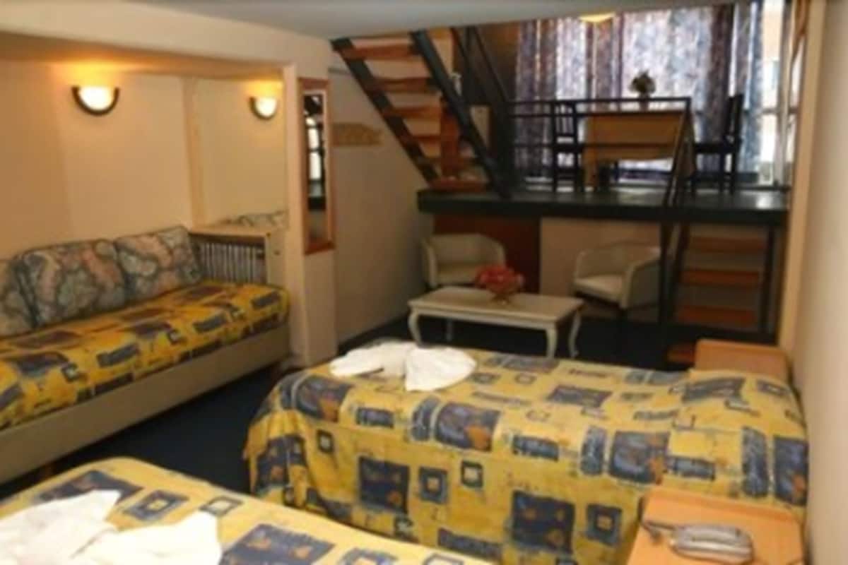 A compact hotel room with two beds, a small table, and a staircase leading to an upper level.