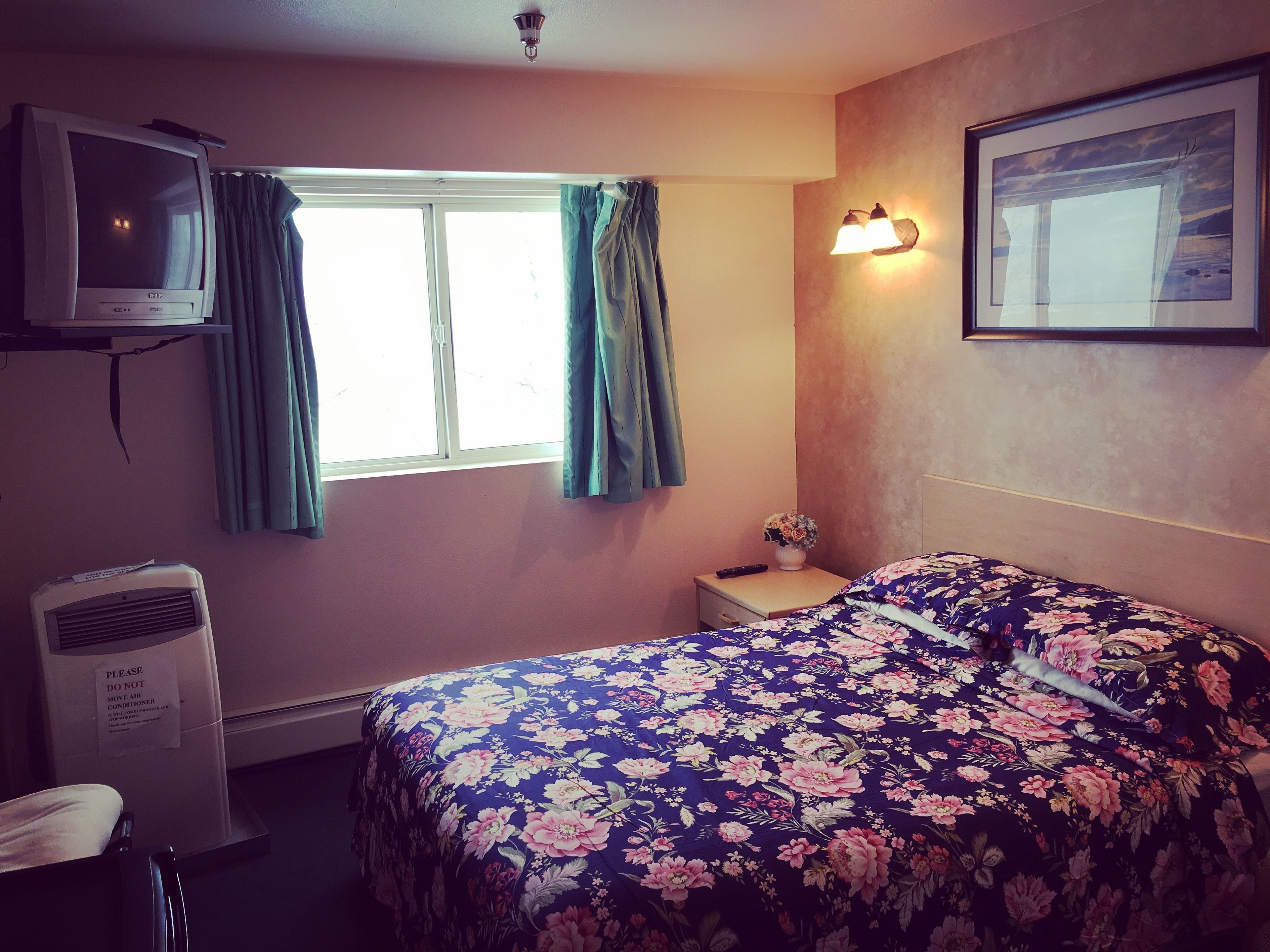A hotel room with a bed, a television, a window with curtains, a nightstand, and a wall-mounted light fixture.