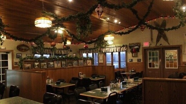 Interior de restaurante con paredes de madera, plantas colgantes e iluminación colgante. Las mesas están dispuestas con sillas y hay cuadros en las paredes.
