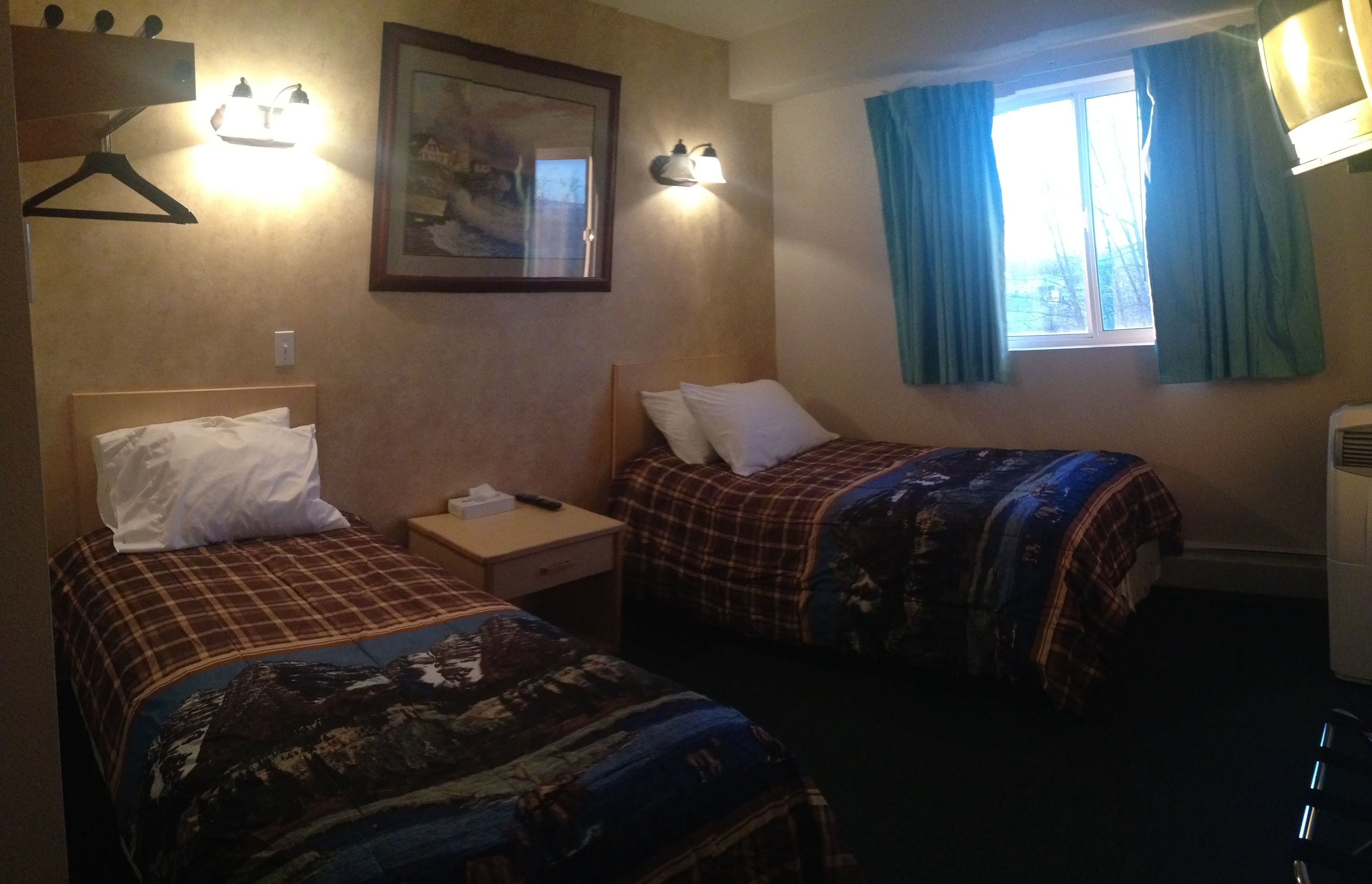 A hotel room with two beds, each with a different patterned bedspread, a bedside table with a lamp, a framed picture on the wall, and a window with curtains.