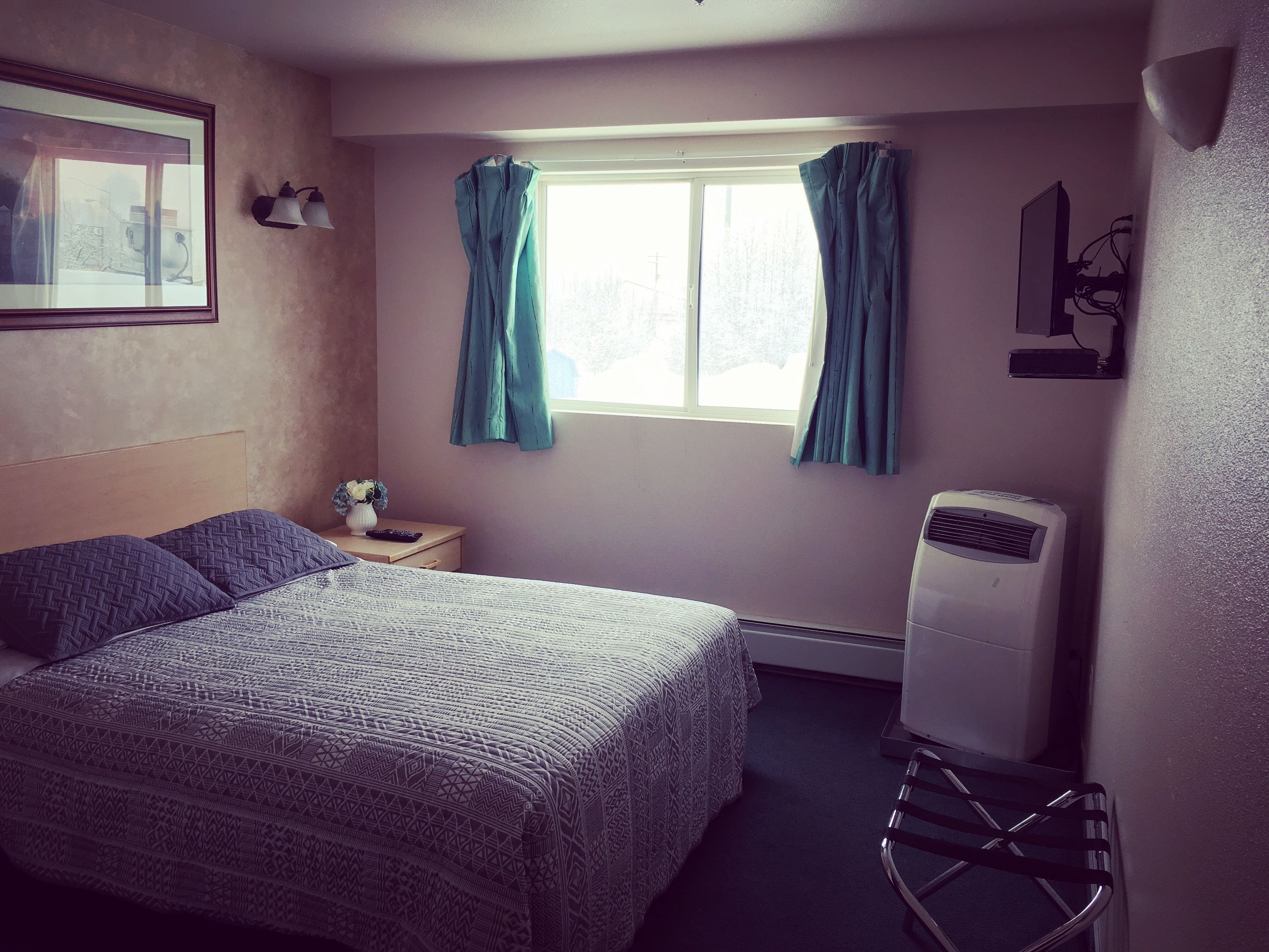 A bedroom with a bed, a nightstand, a window with curtains, a wall-mounted air conditioner, and a framed picture on the wall.