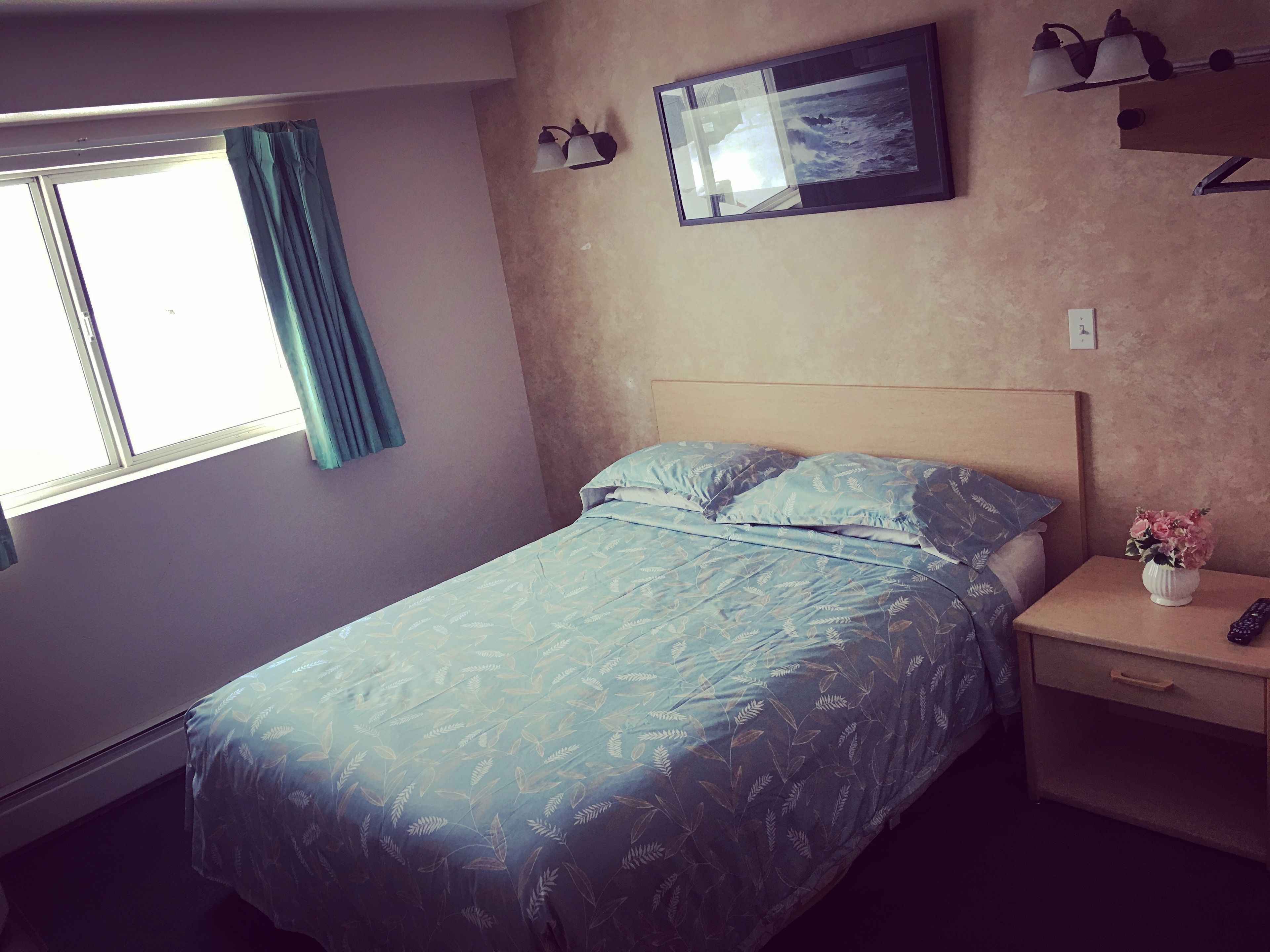 A bedroom with a bed, a nightstand, a vase with flowers, a framed picture, and a window with curtains.