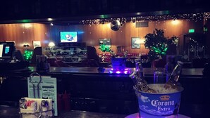 Una zona de bar con una lata de cerveza Corona Extra sobre una mesa iluminada, un monitor de computadora y un árbol de Navidad al fondo.