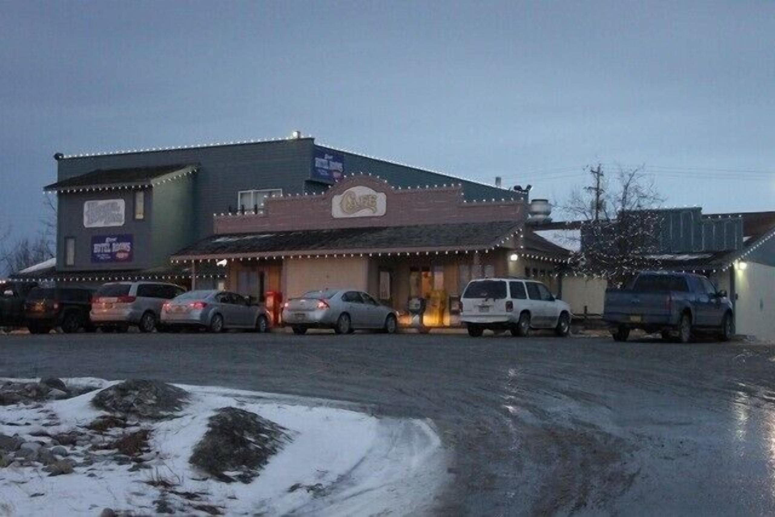Un edificio con un letrero de restaurante, un estacionamiento con varios coches y suelo cubierto de nieve.