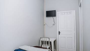Una habitación pequeña y limpia con una cama, una silla, un escritorio y un televisor montado en la pared.