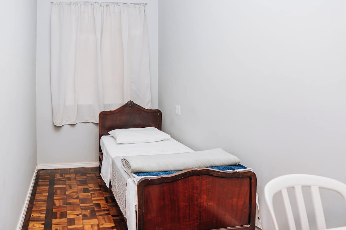A small, neatly made bed with a wooden headboard in a room with a wooden parquet floor and white walls.