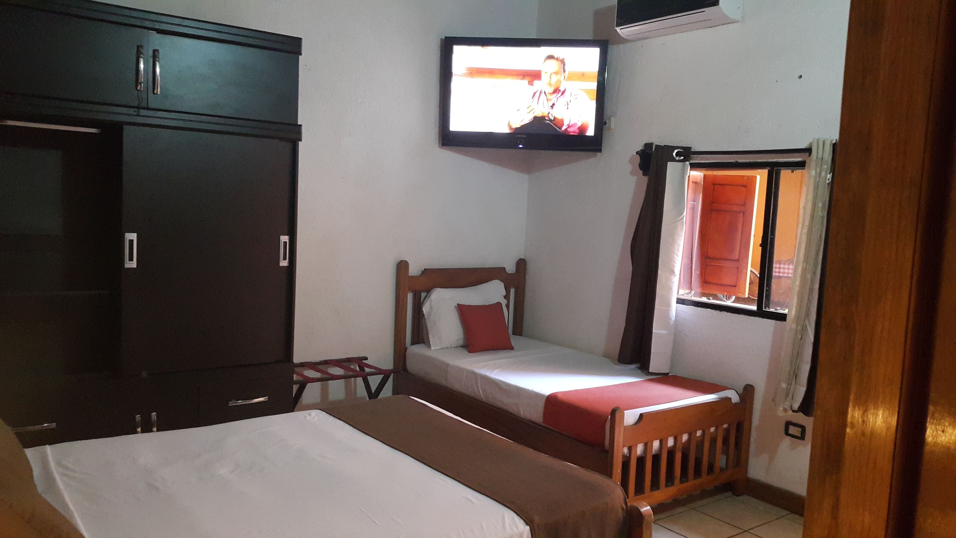 A bedroom with a bed, a wooden wardrobe, a television mounted on the wall, and a window with curtains.