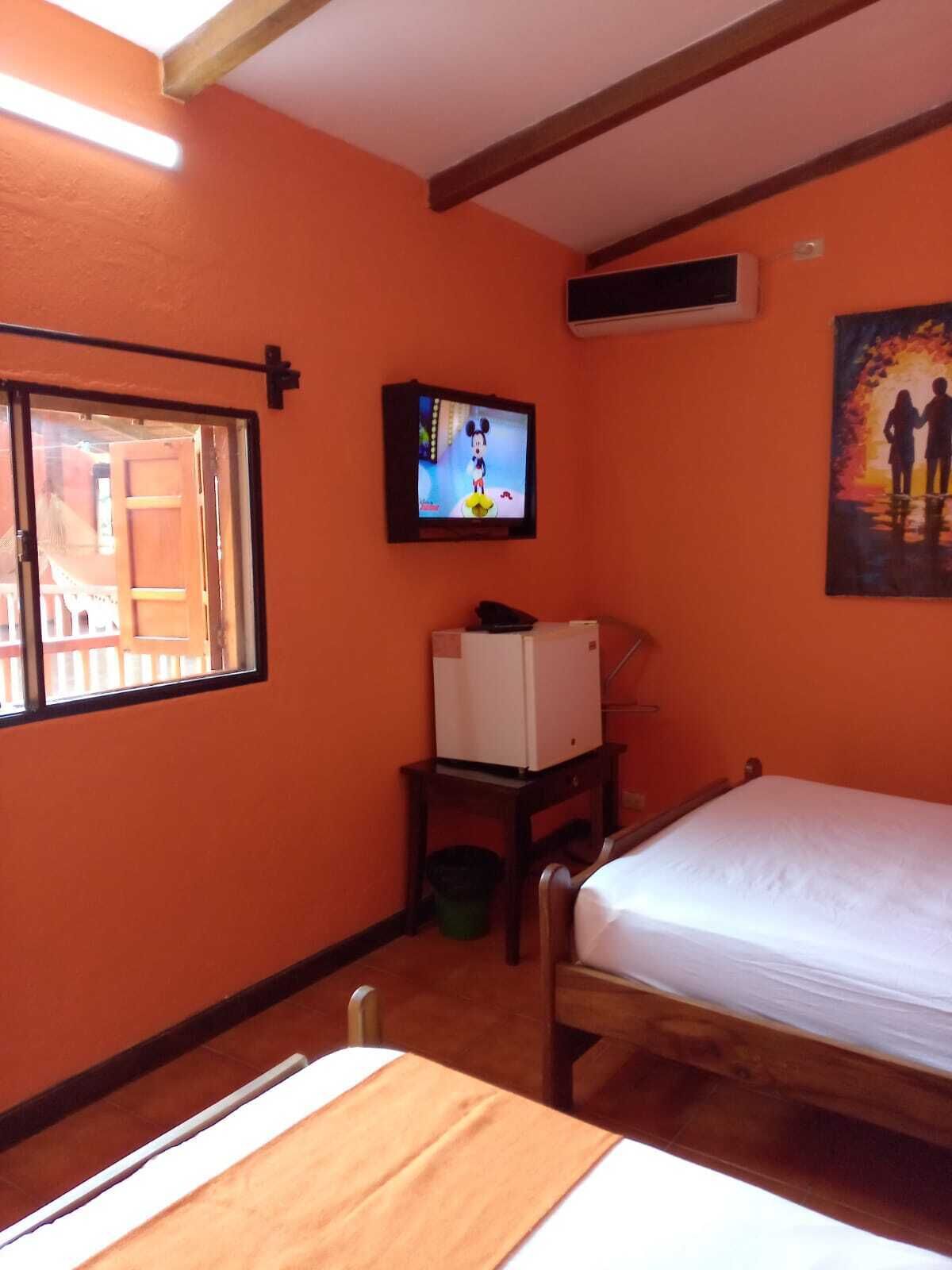 A room with orange walls, a bed, a TV, a small table with a refrigerator, and a painting on the wall.