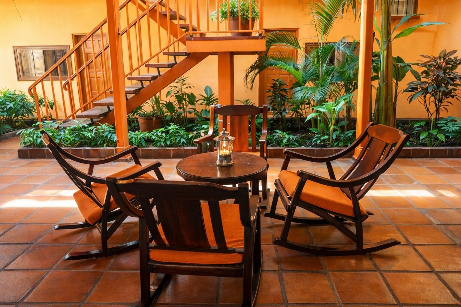 Un espace patio avec une table ronde et quatre fauteuils à bascule, entouré de plantes en pot et d’un escalier.
