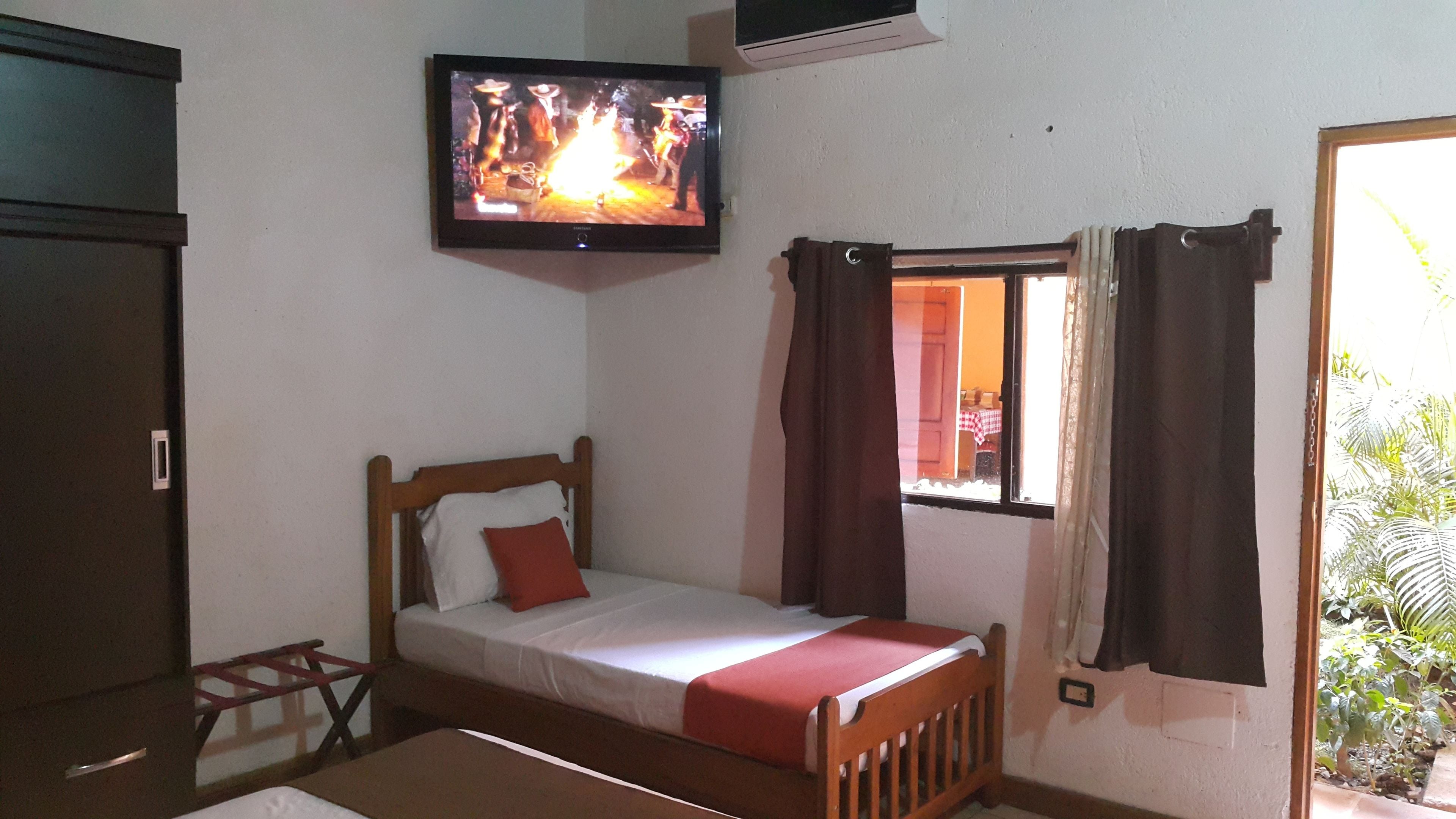 A single bed with a wooden frame, a TV mounted on the wall, and a window with curtains.