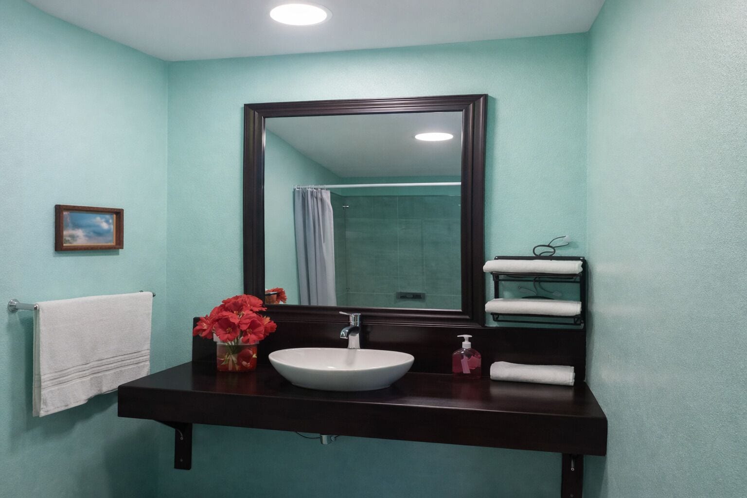 A bathroom with a large mirror, a sink, a towel rack, and a shower visible in the reflection.