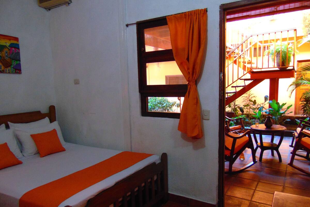A room with a bed, a window with orange curtains, a small table with chairs, and a staircase leading to an outdoor area with plants.