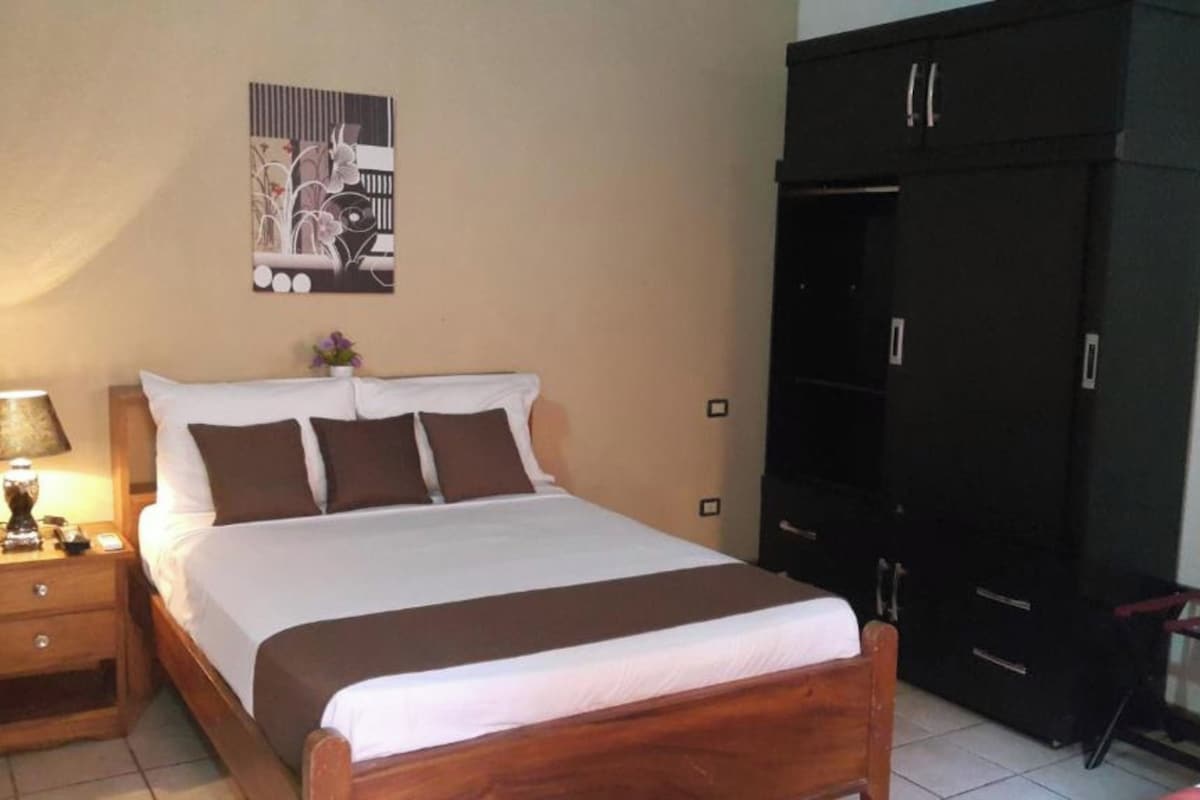 Un dormitorio con cama, mesita de noche, lámpara, armario y un cuadro en la pared.