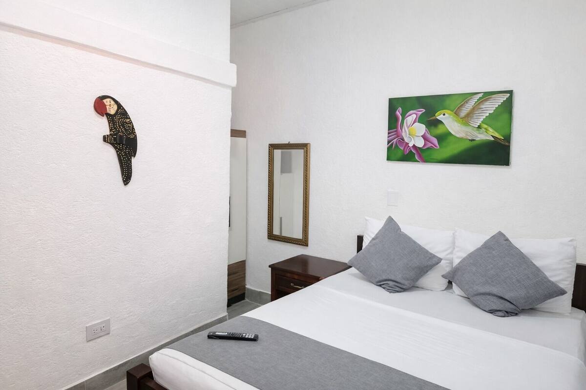 A hotel room with a bed, pillows, a remote control, a wall-mounted mirror, and a painting of a bird and flower.