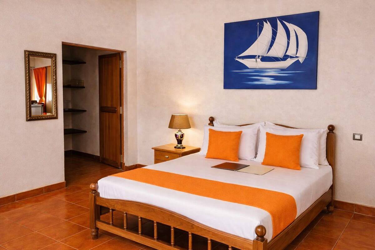 A hotel room with a wooden bed, an orange throw, a nightstand with a lamp, a mirror, and a painting of a sailboat.