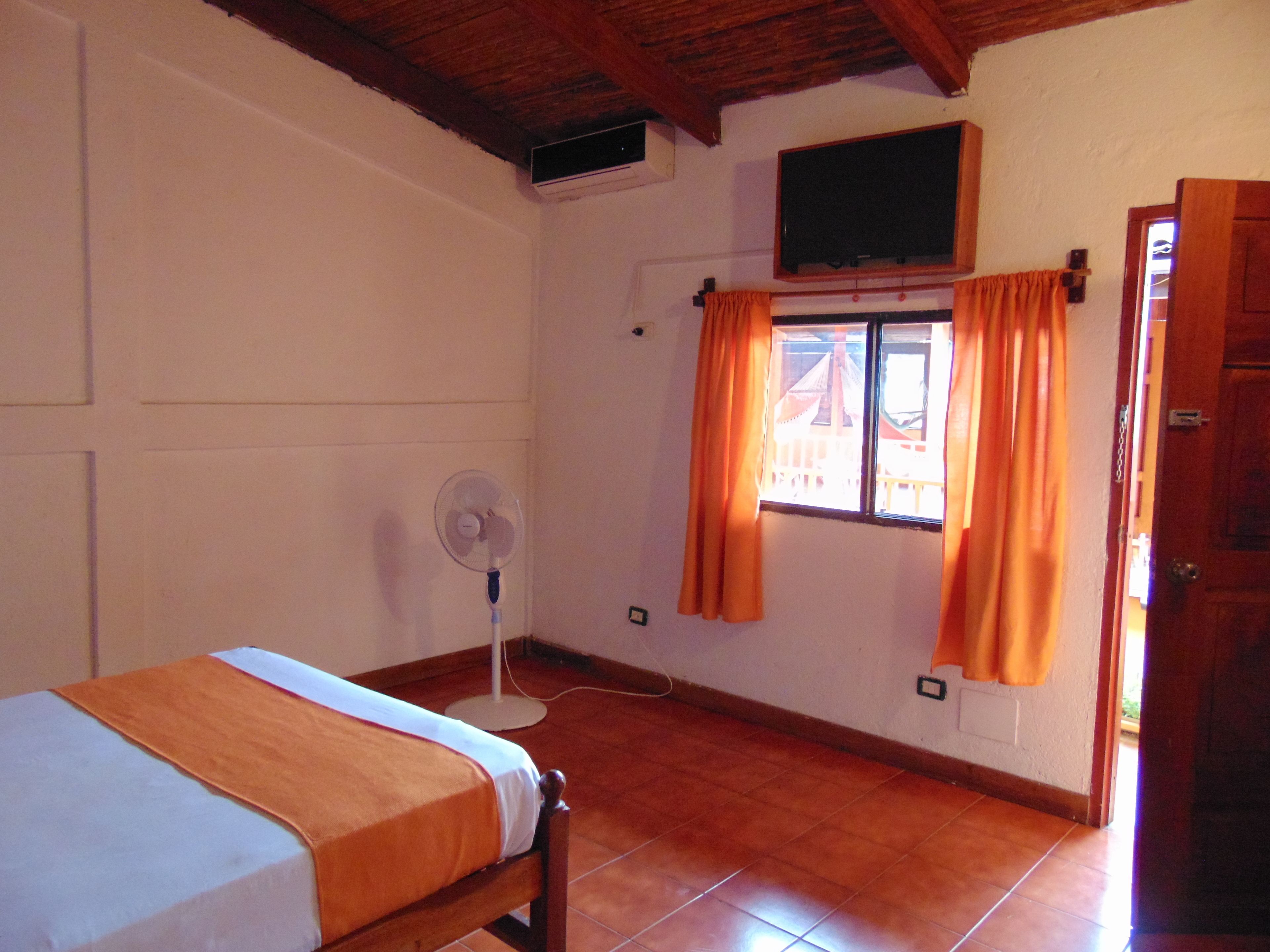 A room with a bed, a standing fan, a window with curtains, and a television mounted on the wall.