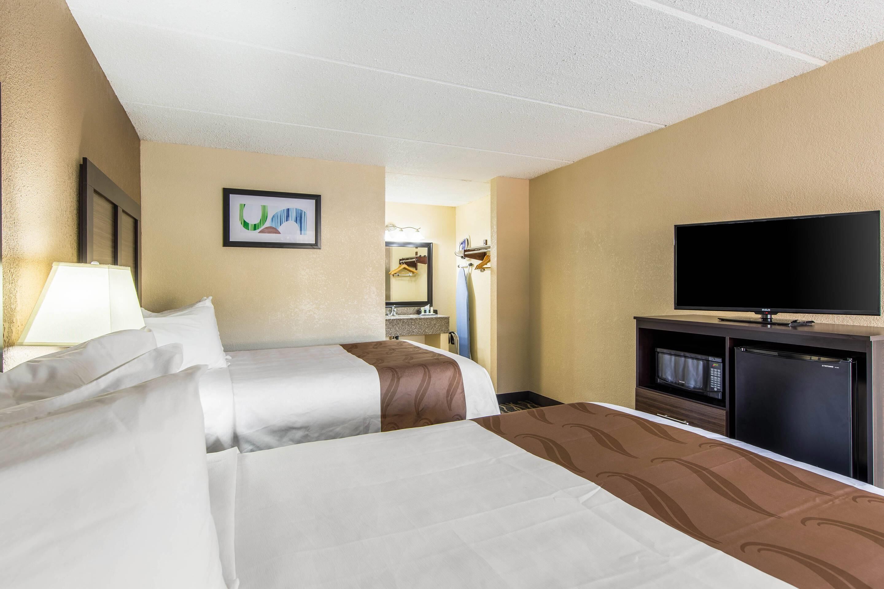 A hotel room with two beds, a television, a desk with a microwave, and a bathroom visible in the background.