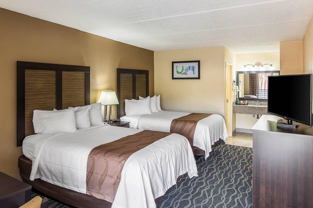 Quarto de hotel com duas camas, uma escrivaninha, televisão e banheiro visível através de uma porta aberta.