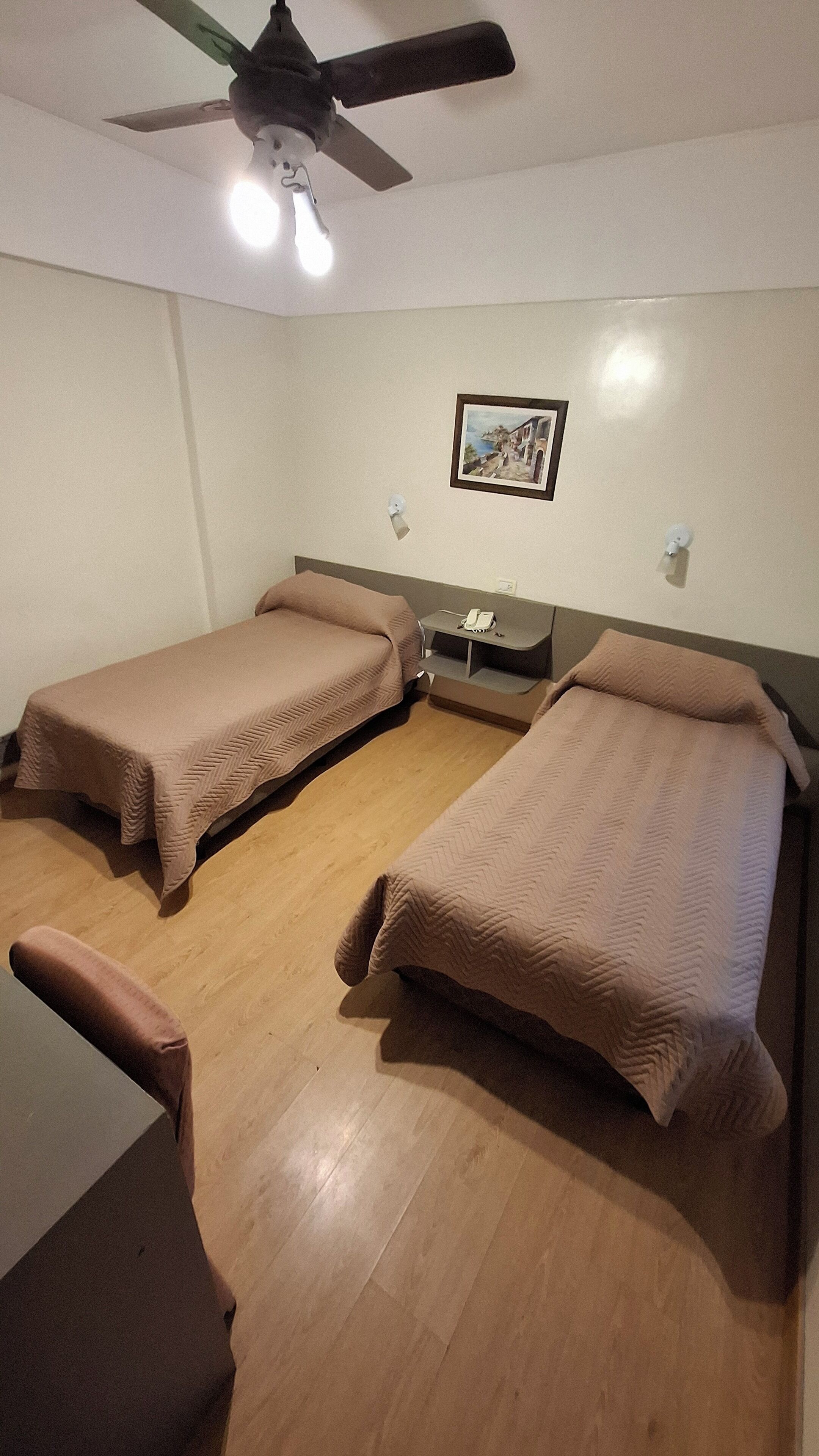 Ein Zimmer mit zwei Einzelbetten, einem Deckenventilator, einem Gemälde an der Wand und einem kleinen Tisch mit Telefon.