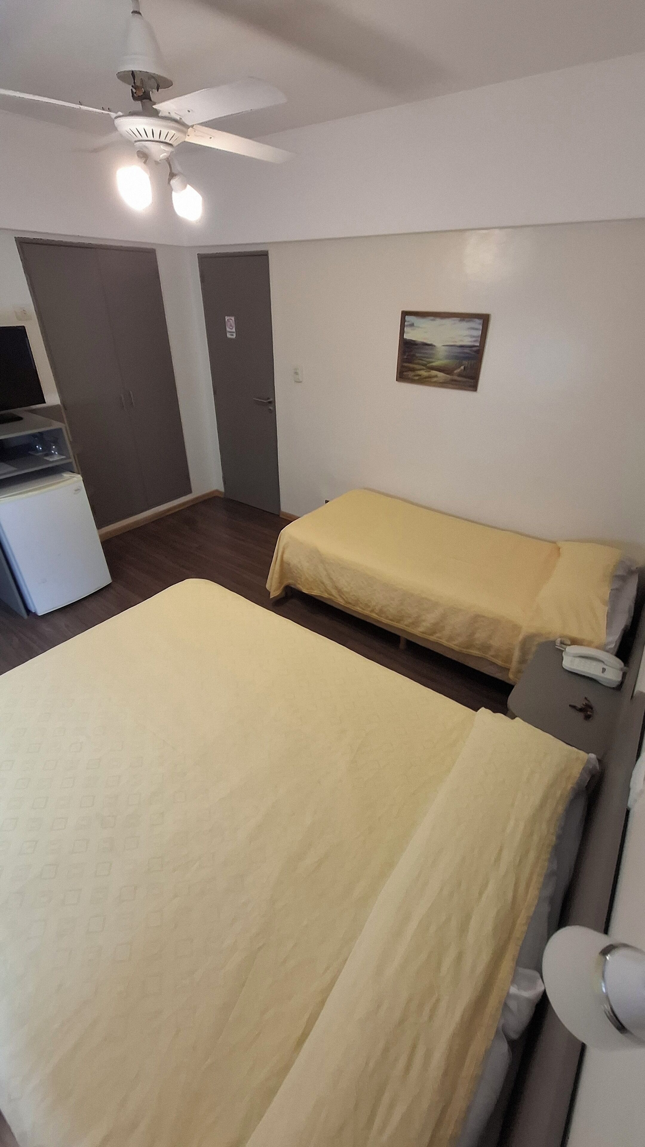 Una habitación con dos camas individuales, un ventilador de techo, un refrigerador pequeño y un cuadro en la pared.