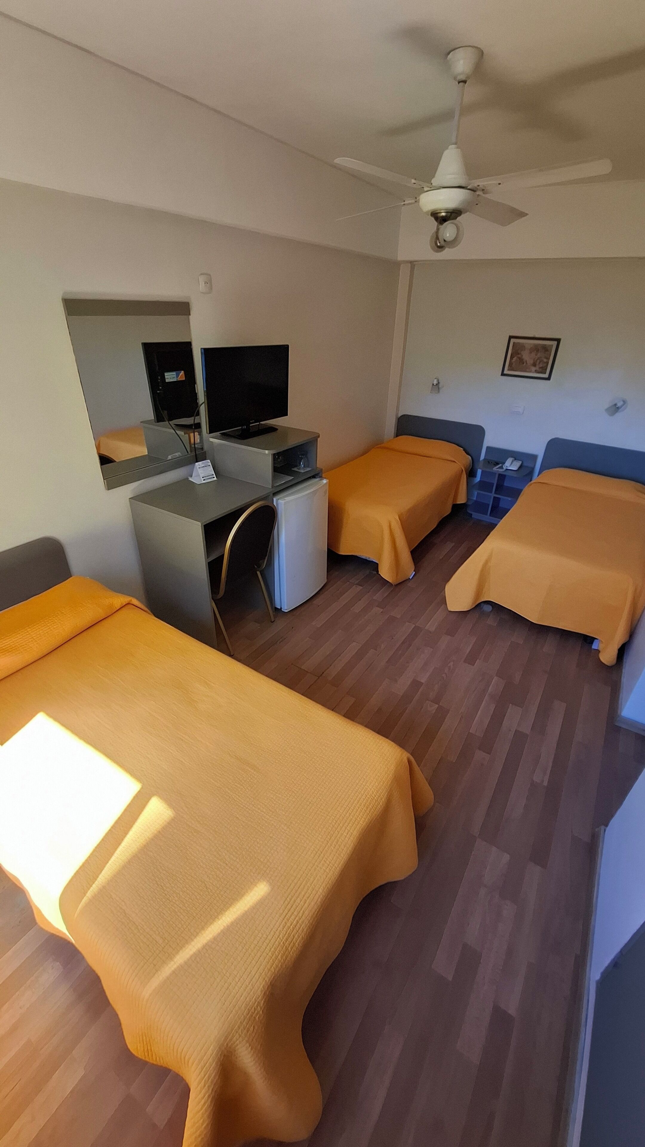 Habitación de hotel con dos camas individuales, un escritorio con computadora, un refrigerador pequeño y un ventilador de techo.