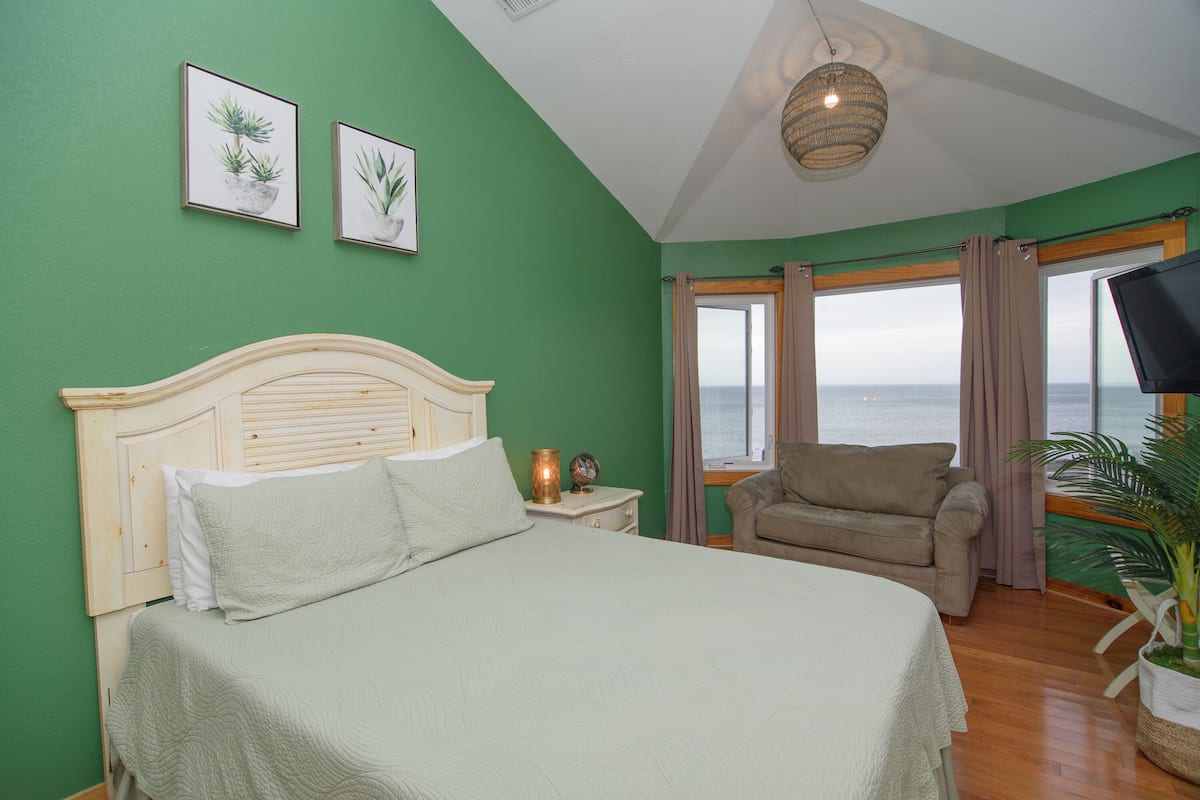 A bedroom with a bed, a nightstand, a chair, a television, and a view of the sea through the window.