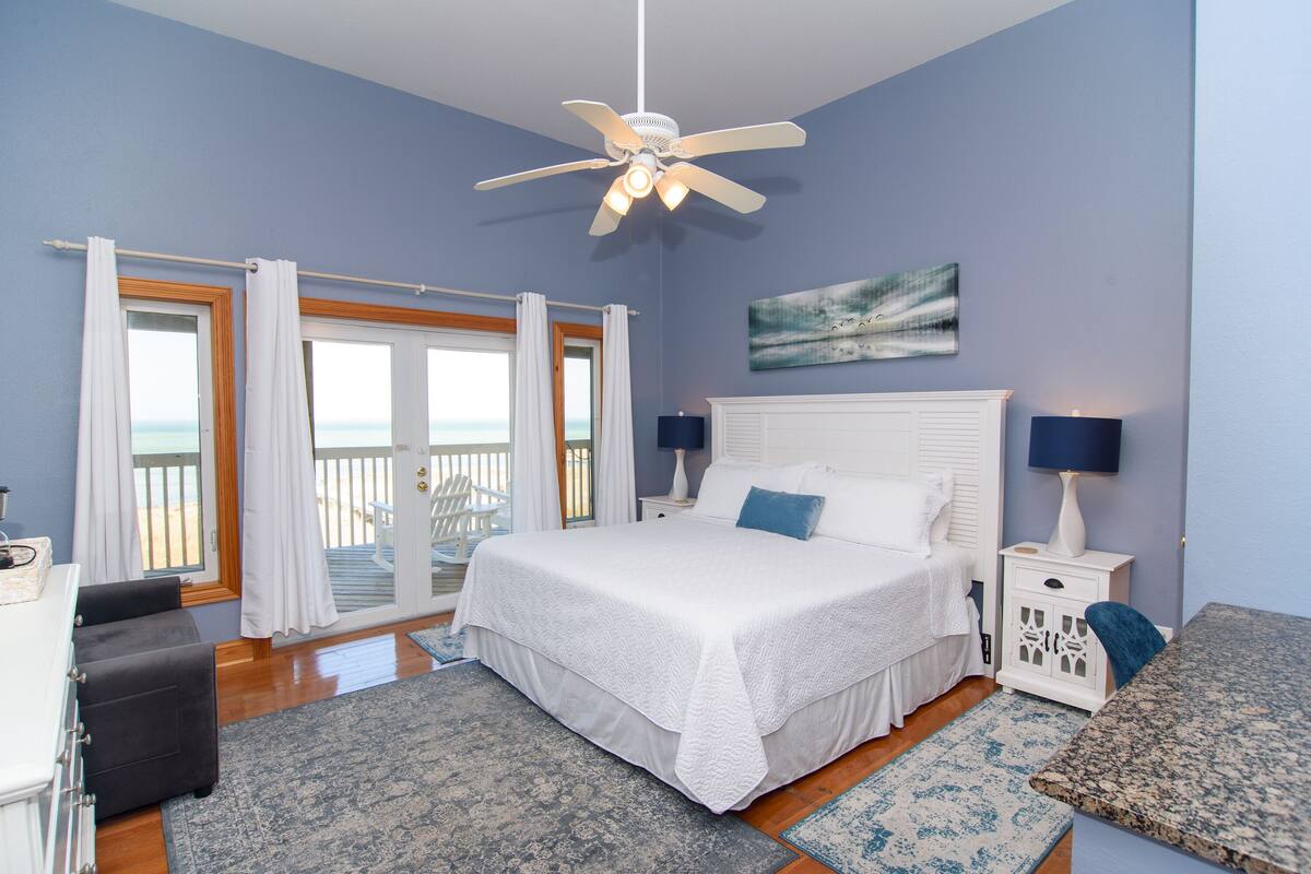 A bedroom with a bed, a ceiling fan, a nightstand with a lamp, a painting on the wall, and a view of the ocean through a sliding door.