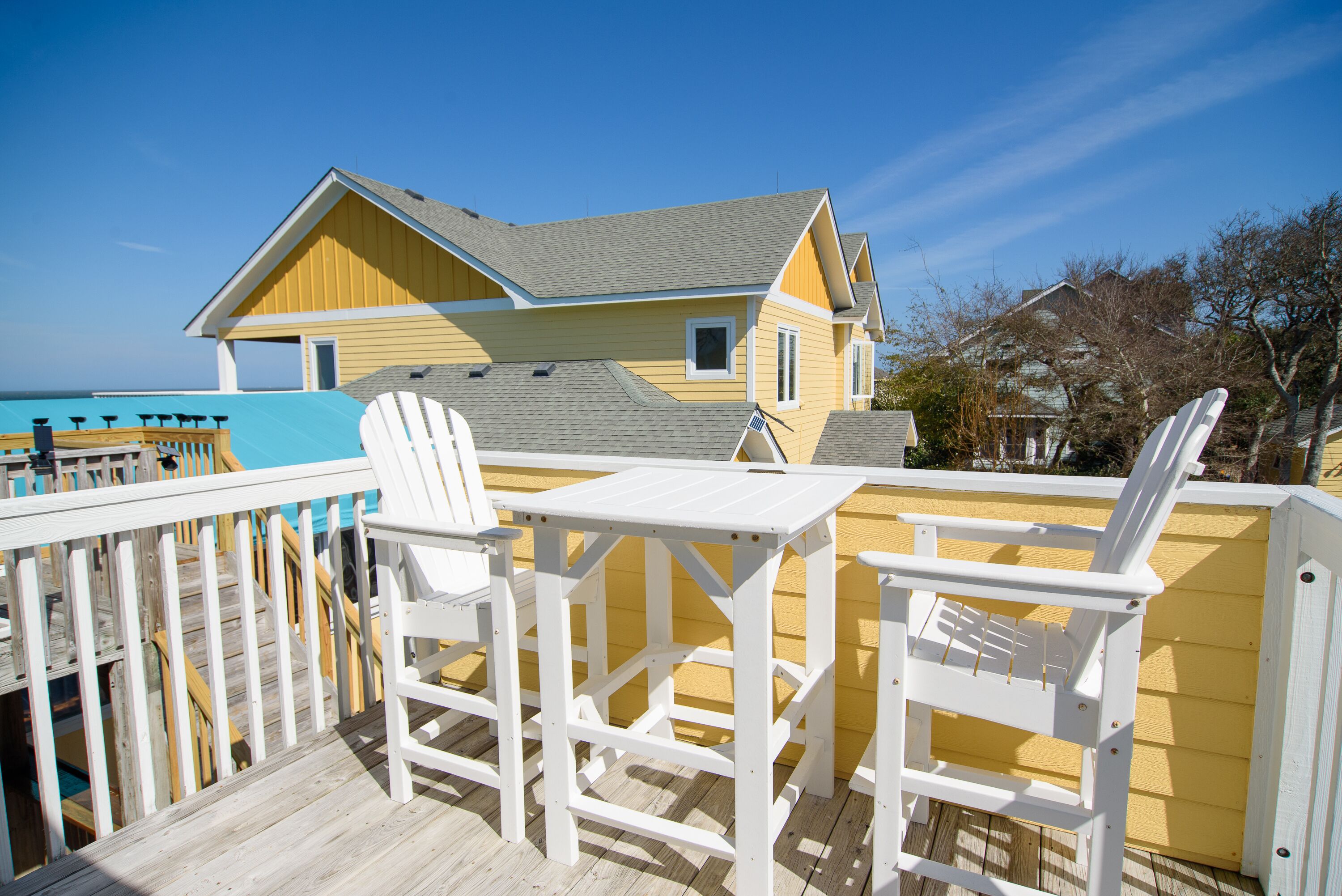 Eine Holzterrasse mit weißen Stühlen und einem Tisch, mit Blick auf einen Strand und ein gelbes Haus mit grauem Dach.